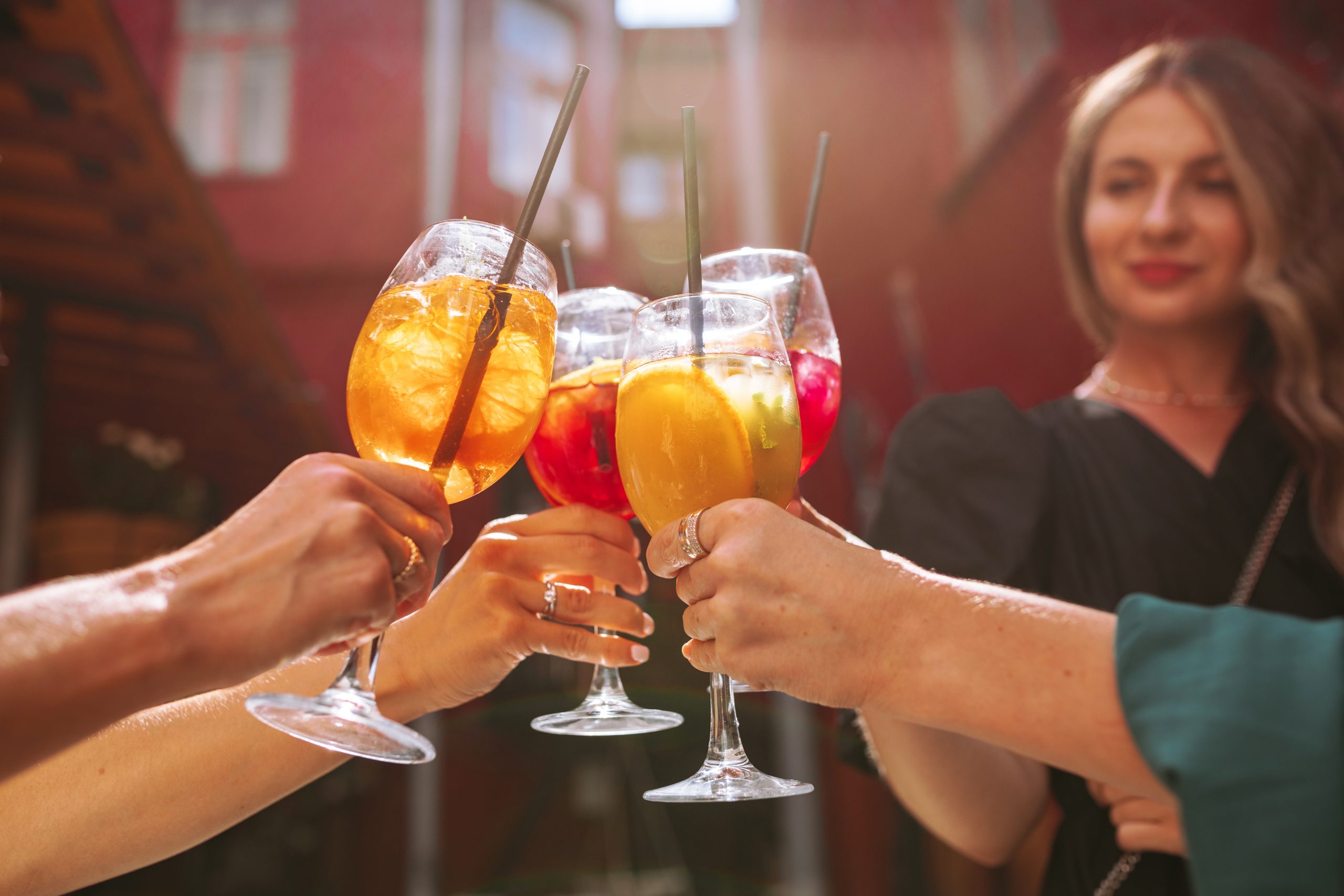 Vier Hände stoßen mit bunten Aperitif-Gläsern an, im Hintergrund lächelt eine Frau vor einer roten Hauswand