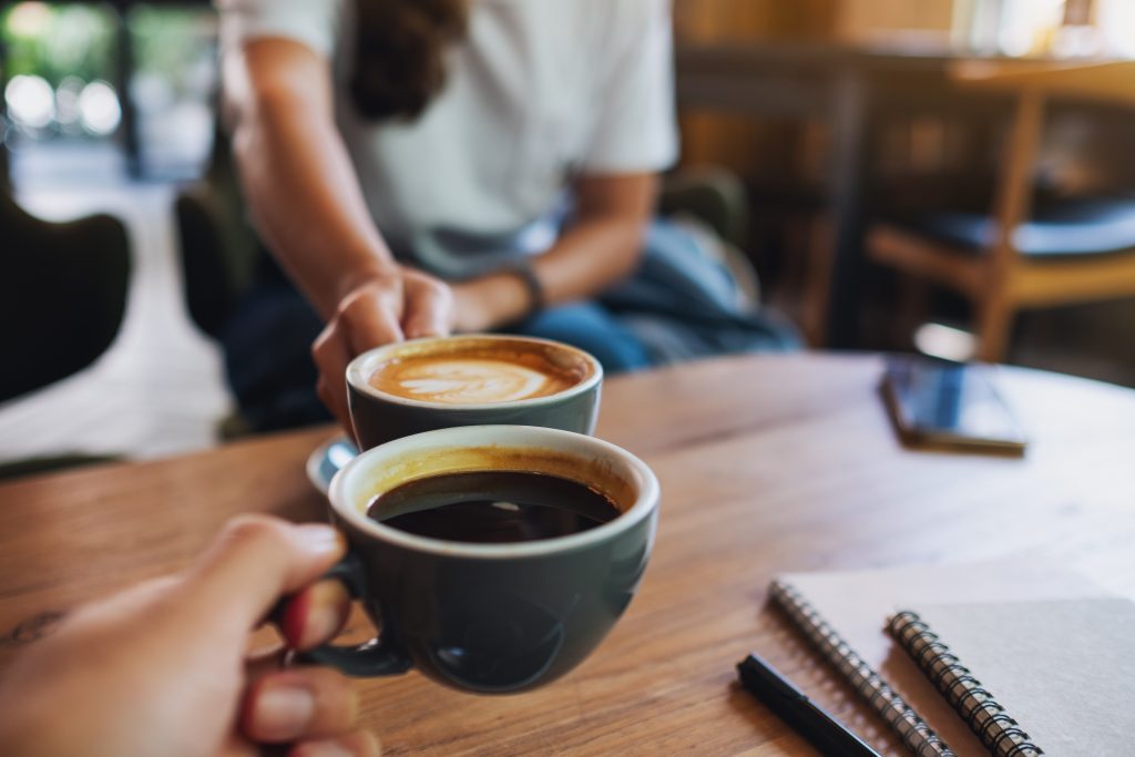 Zwei Personen stoßen mit Tassen Cappuccino und schwarzem Kaffee an, daneben liegen Notizbücher und ein Smartphone