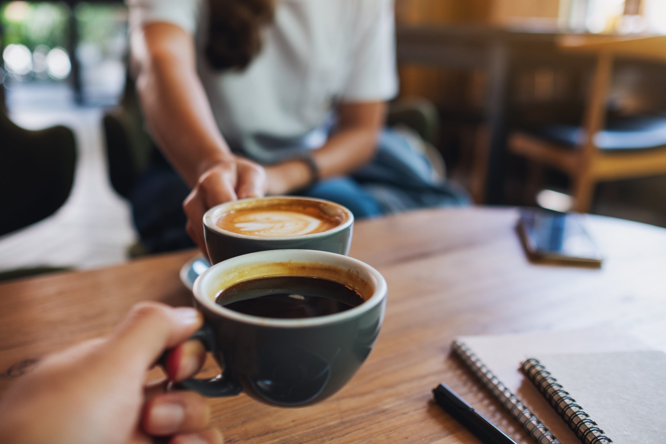 Zwei Personen stoßen mit Tassen Cappuccino und schwarzem Kaffee an, daneben liegen Notizbücher und ein Smartphone