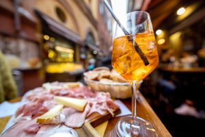 Aperol Spritz mit Eiswürfeln im Glas, daneben eine Antipasti-Platte mit Schinken, Käse und Brot