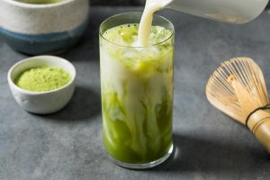 Milch wird in ein Glas mit Eiswürfeln und Matcha gegossen, daneben Matchapulver und ein Bambusbesen
