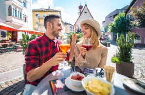 Lachendes Paar sitzt beim Aperitif mit Aperol Spritz und Rotwein an einem Tisch auf einem Platz mit Kirche, vor ihnen Chips und Snacks