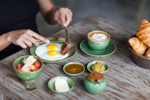 Frühstück mit Spiegelei, Speck, Cappuccino, Obstschale und Croissants auf einem Holztisch