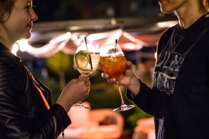 Zwei Personen stoßen abends im Freien mit Gin Tonic und Aperol Spritz an, im Hintergrund warmes Licht